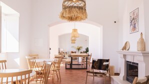 A spacious living room with a fireplace, a dining area with wooden chairs, and a large window allowing natural light. - Quinta da Baleeira (Tavira)