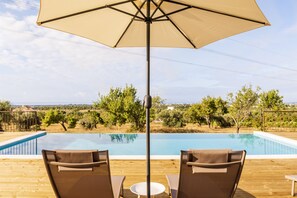 Zona de piscina con terraza de madera, dos sillones, una mesa y una sombrilla.