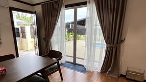 Een kamer met een houten eettafel, stoelen, een bank en een glazen schuifdeur die uitkomt op een buitenruimte met uitzicht op groen en een gebouw.