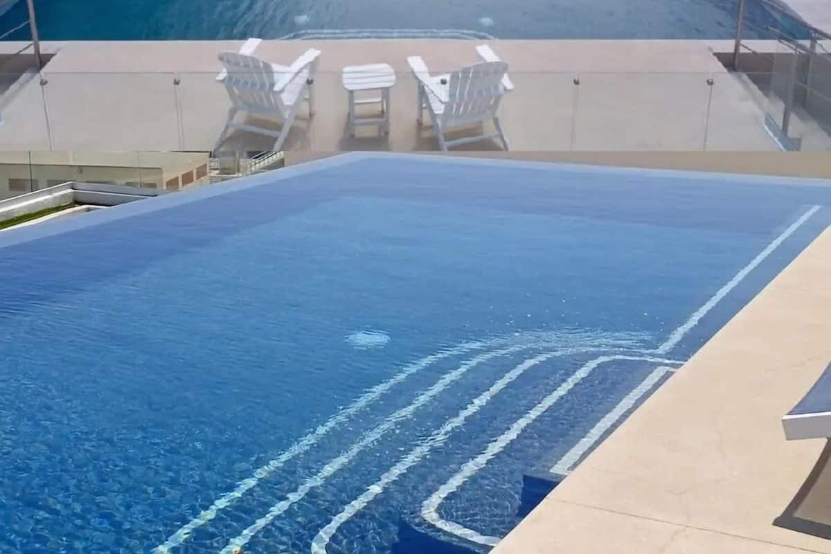 A rooftop infinity pool with clear blue water, white lounge chairs, and a view of the ocean and distant mountains.
