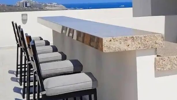 A rooftop terrace with a long row of chairs and a small bar area, overlooking a coastal view.