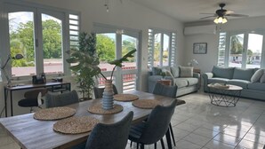 Ein geräumiger Wohnbereich mit Esstisch, Stühlen und einer Couch. Große Fenster sorgen für Tageslicht und Blick auf die grüne Umgebung.
