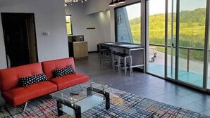 Un salón moderno con un sofá rojo, una mesa de centro de cristal y vistas a un paisaje verde a través de ventanas amplias.