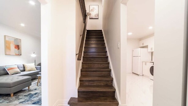 A staircase leading to a second floor, a gray sofa with a patterned rug, and a kitchen area with white cabinets and appliances.