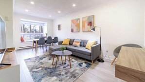A modern living room with a grey sofa, a coffee table, and a dining area with chairs. There are abstract paintings on the wall and a window providing natural light.