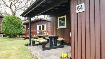 En træhytte med nummer 64 på væggen, en overdækket terrasse med et bord og bænke, og en veltrimmet busk.