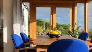 A dining area with a wooden table and blue chairs, a bowl of fruit, and a view of greenery through large windows. - 3-bed Cottage Near Lake Vyrnwy, Garden & Petsok (Oswestry)