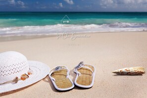 A beach scene with a white hat, flip-flops, and a seashell on the sand, with a clear view of the ocean and a logo in the sky.