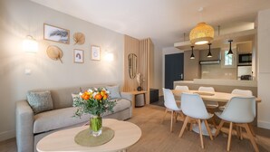 Una sala de estar moderna con un sofá, una mesa de comedor con sillas, un florero, luces fijadas a la pared y una zona de cocina al fondo.
