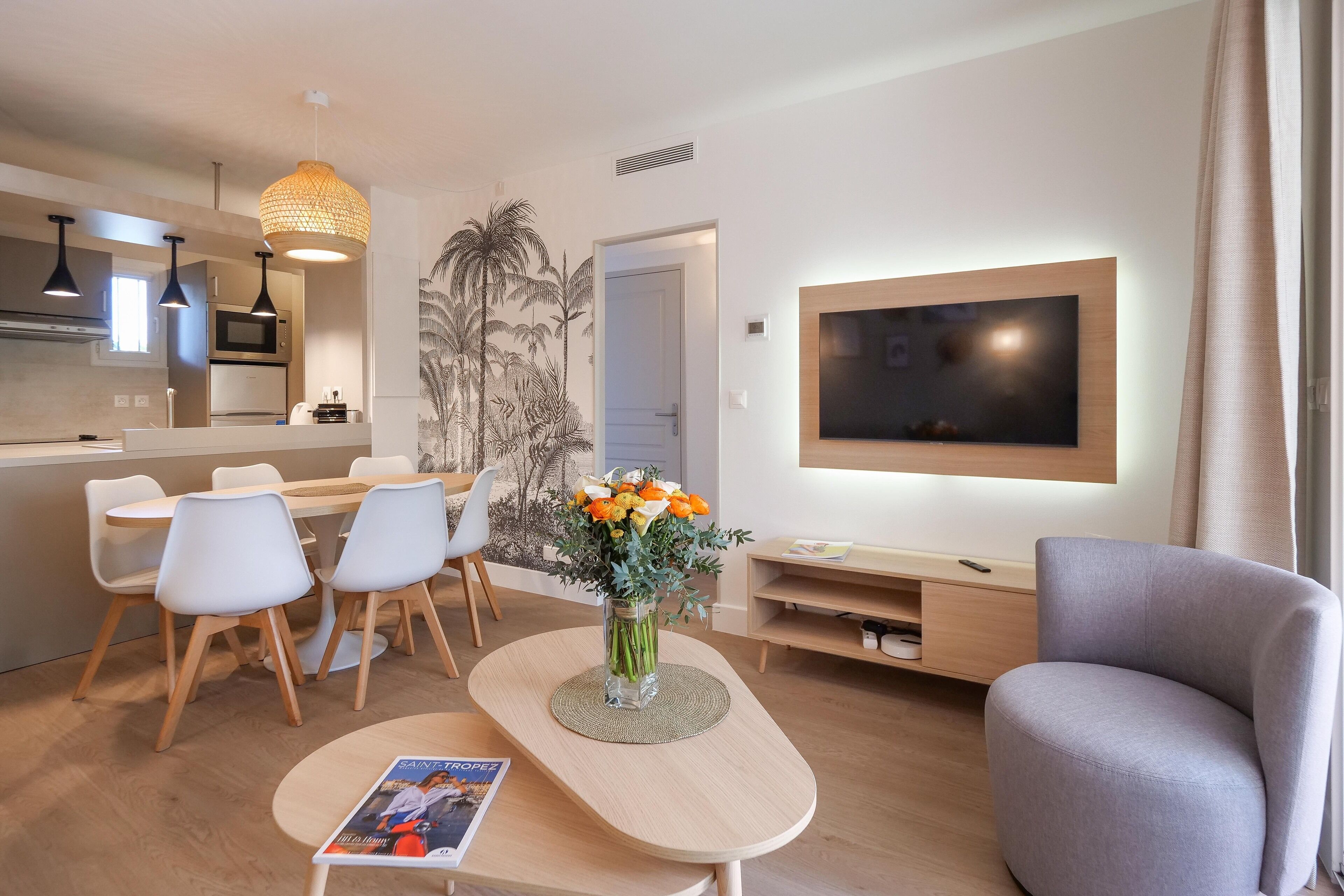 Een moderne woonkamer met een flatscreen televisie, een grijze fauteuil, een eethoek met witte stoelen en een keuken met een magnetron.