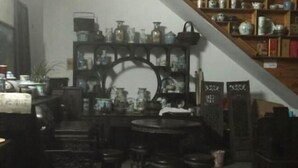 Una habitación tenuemente iluminada con una mesa redonda y varios taburetes, rodeada de estanterías llenas de objetos de cerámica.