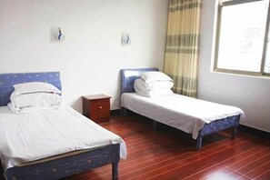 Une chambre avec deux lits simples, une petite table de chevet en bois, une fenêtre avec un rideau et un luminaire fixé au mur.