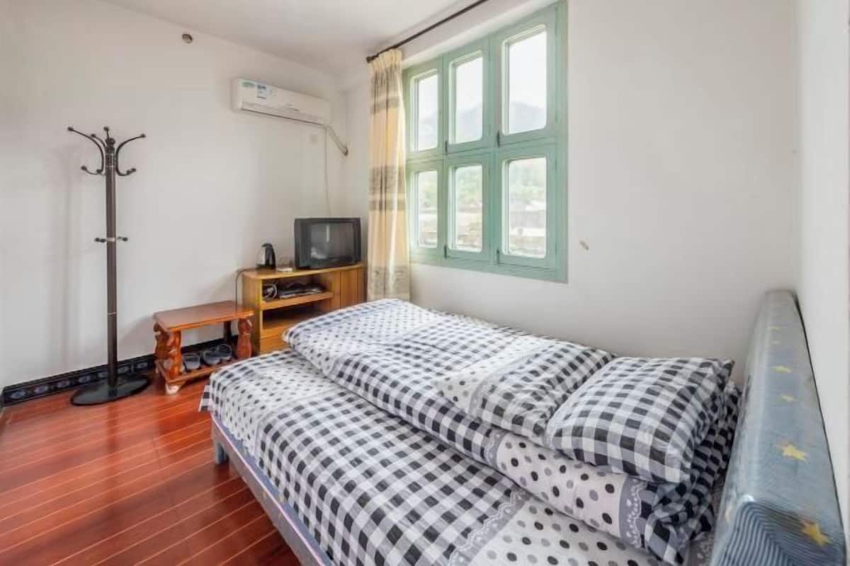 A small, tidy room with a bed, a television, a coat rack, and a window with curtains.