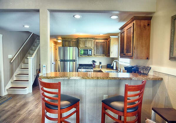 A kitchen with wooden cabinets, a stainless steel refrigerator, a microwave, and a granite countertop. There are two wooden bar stools with black seats. - Treetop River Cabins (Center Point)