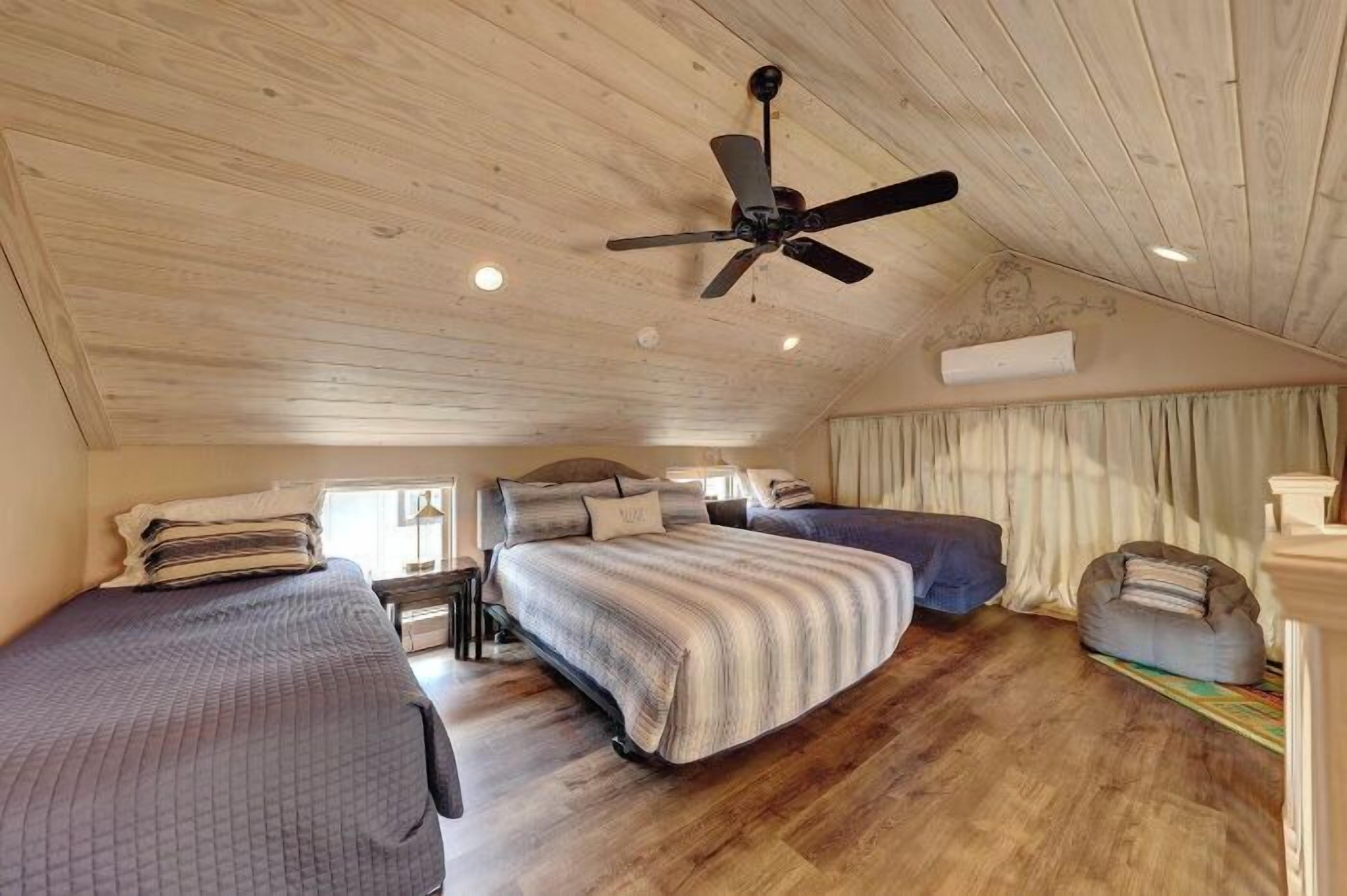 A cozy bedroom with a sloped ceiling, a ceiling fan, two beds, a bean bag, and a window with curtains.