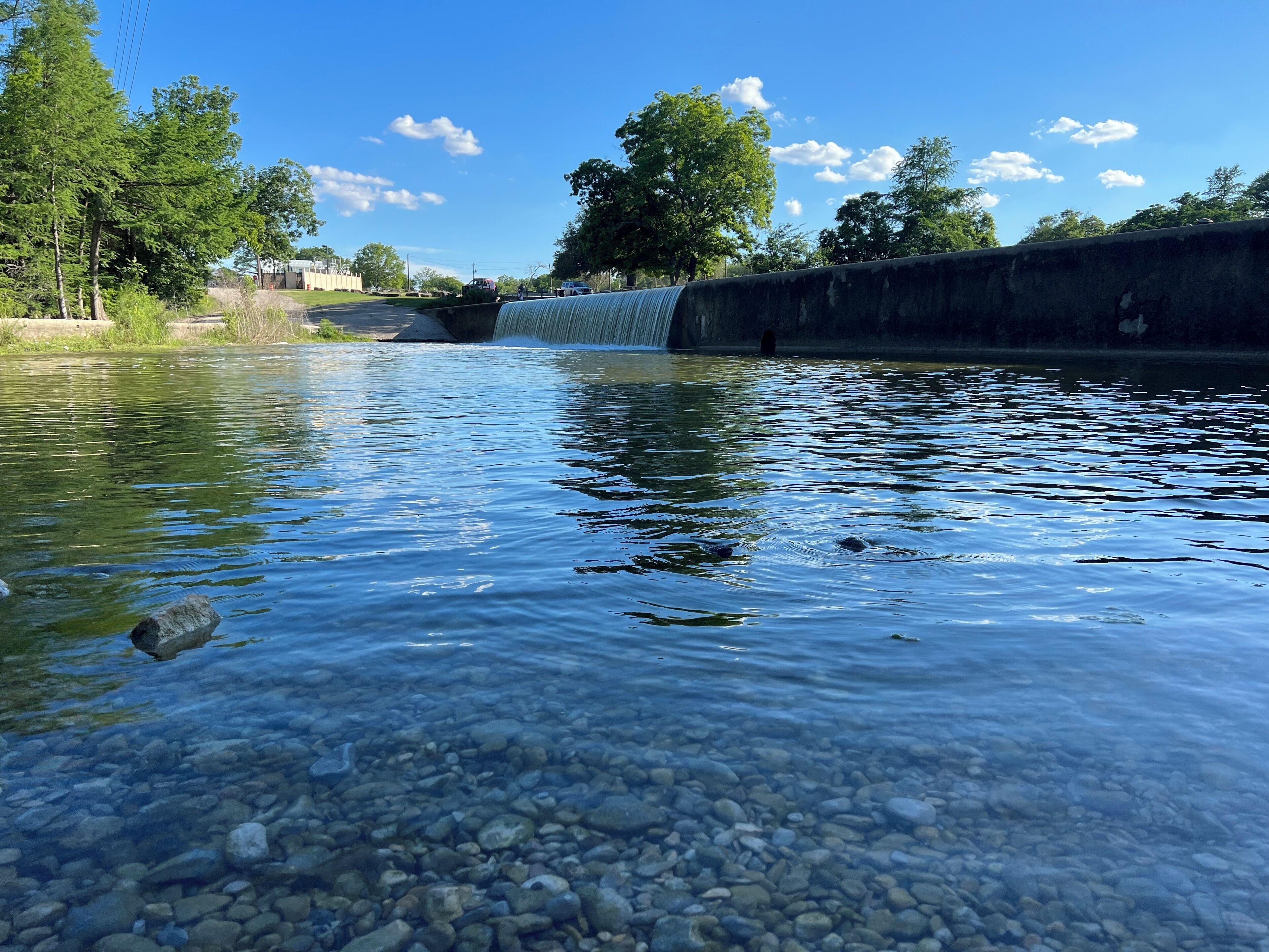 A clear river with a concrete bridge, pebbles at the water's edge, and trees in the background.
