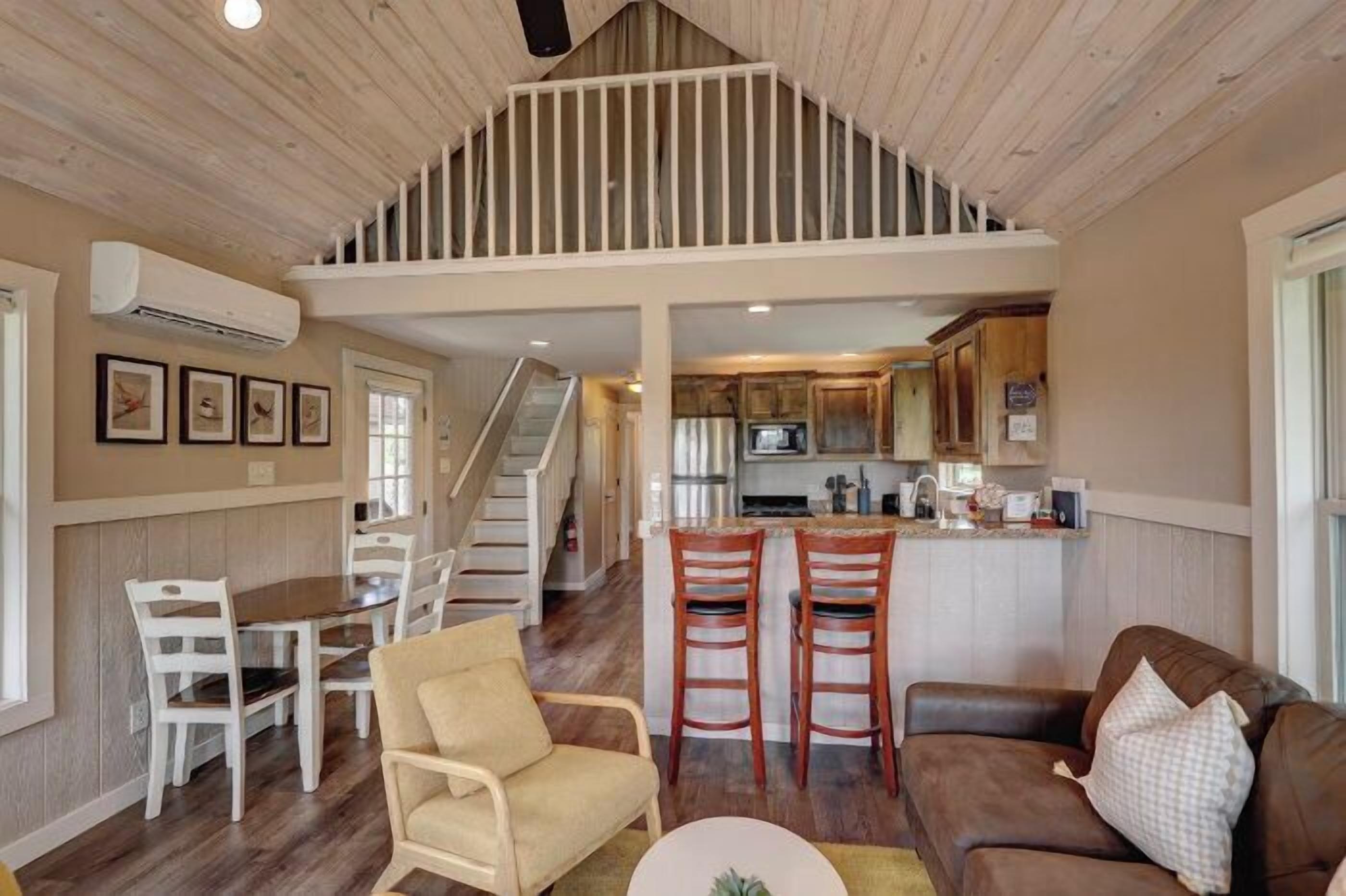 A spacious living area with a high ceiling, wooden flooring, a dining table with chairs, a sofa, and a kitchen area with a bar counter.