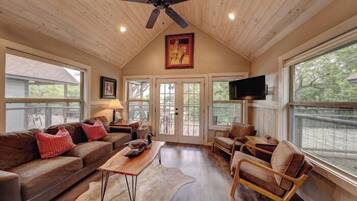 A cozy living room with a wooden ceiling, a ceiling fan, a large window, a television, a brown leather sofa, a wooden coffee table, and a brown leather armchair.