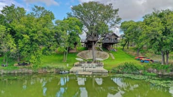 A lakeside view with a wooden house on stilts, surrounded by lush greenery and a calm pond.