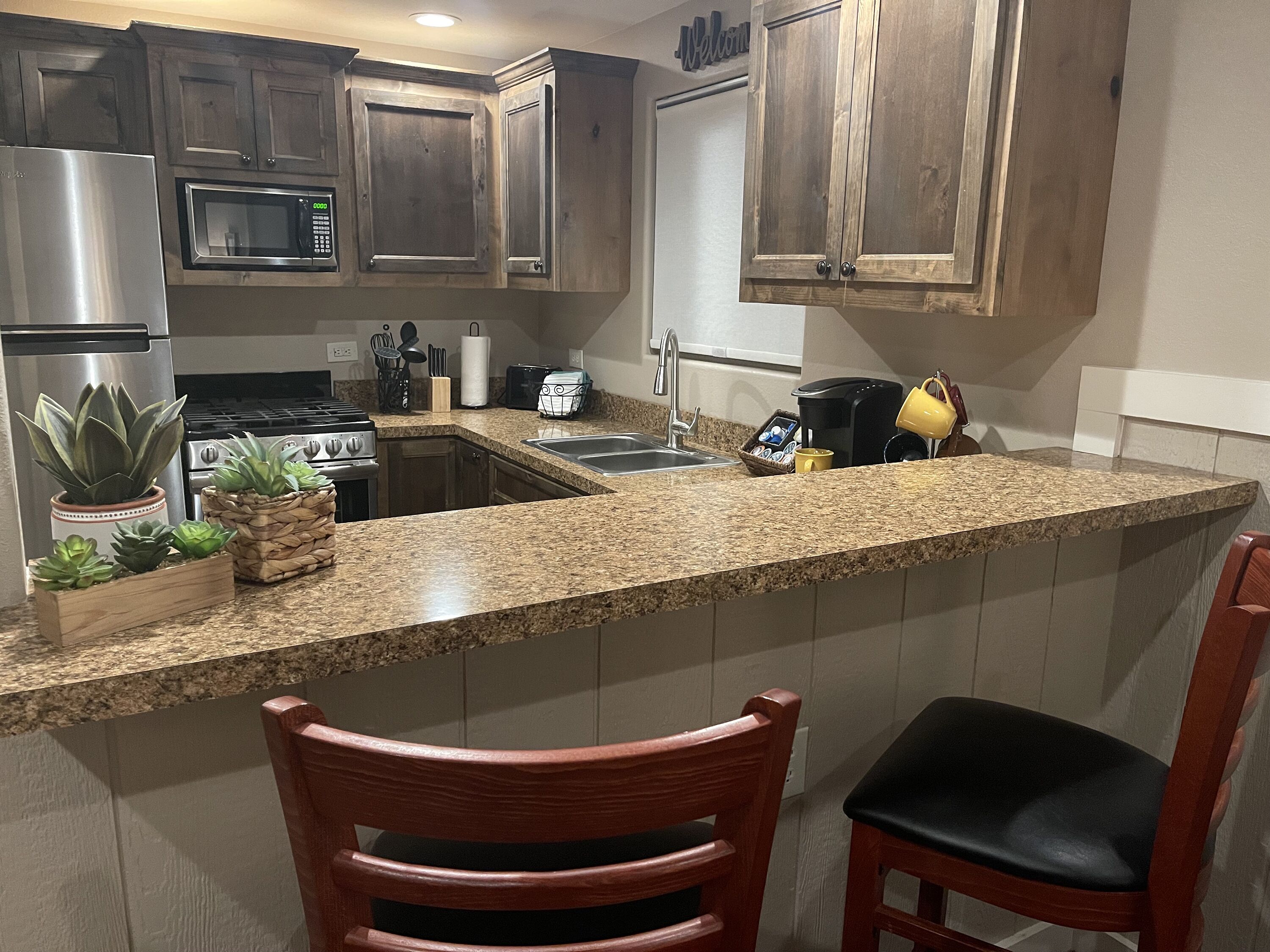 A kitchen with granite countertops, wooden cabinets, a stainless steel refrigerator, a microwave, a stove, and a coffee maker. There are two chairs and a potted plant.