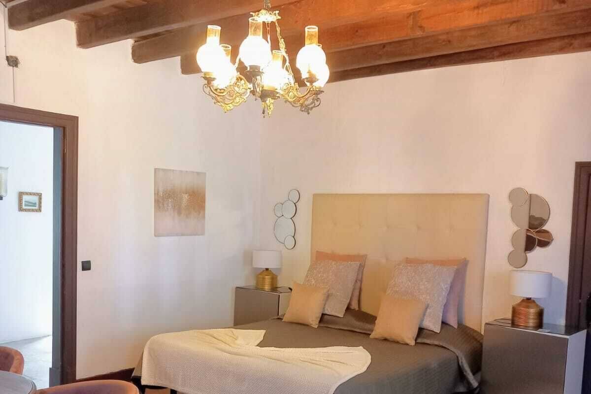 A bedroom with a wooden ceiling, a chandelier, a bed with multiple pillows, a bedside table with a lamp, and a chair.