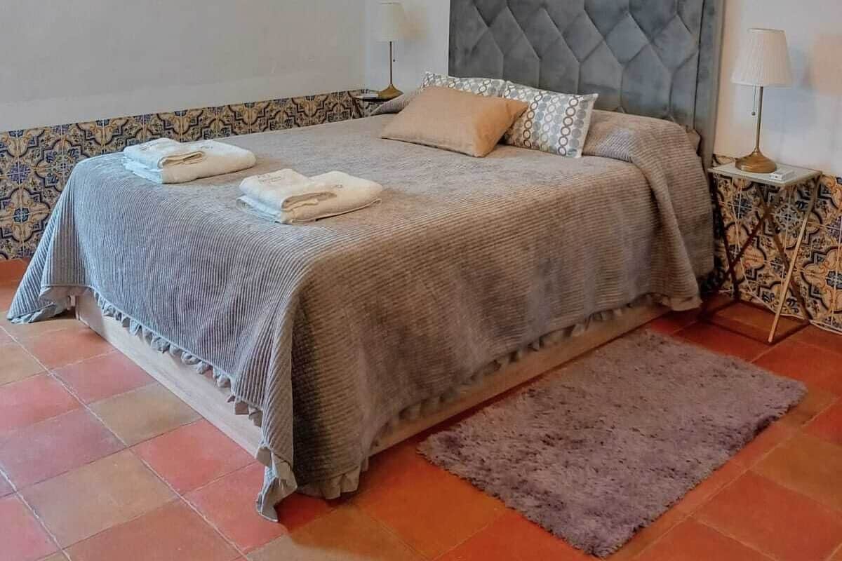 A neatly made bed with a grey headboard, a bedside lamp, and a patterned rug on a tiled floor.