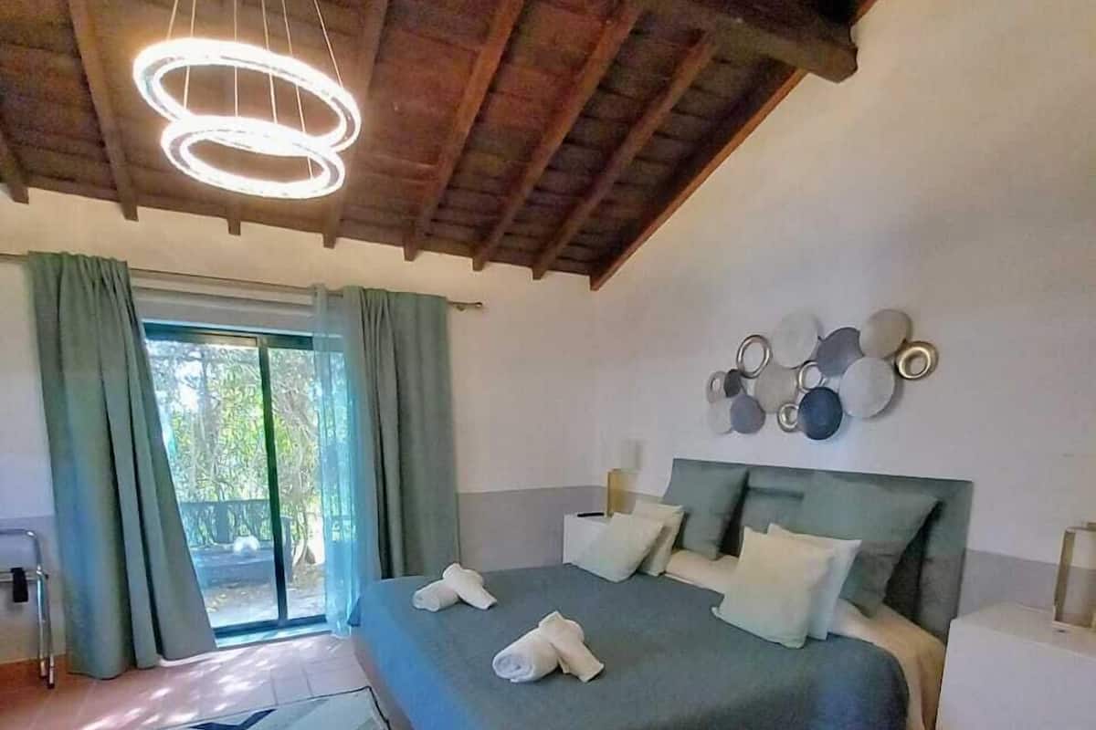 A bedroom with a bed, a nightstand, a wall-mounted light fixture, and a view of the outdoors through a window.