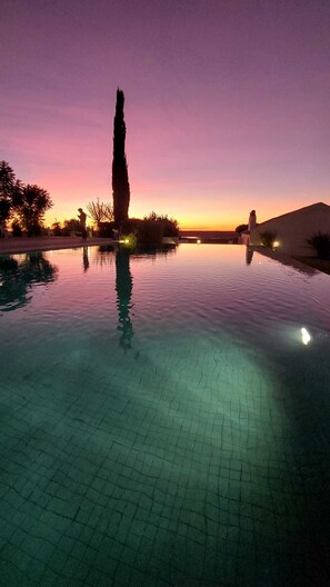 An infinity pool with a clear view of the sunset, a tall tree, and a building in the background. - Agroturismo Herdade Fonte Ferreira (Avis)