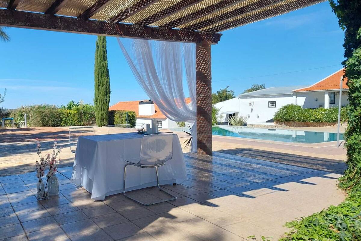 A covered outdoor area with a table set for an event, a pool, and a white building in the background.
