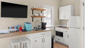 A modern kitchen with white cabinetry, a built-in oven, a microwave, and a coffee maker. There are two white stools and a small sink. - Hanby Hideaway~ Cottages @ Kure ~ Beach, Fishing, Pool, Walk to Pier (Kure Beach)