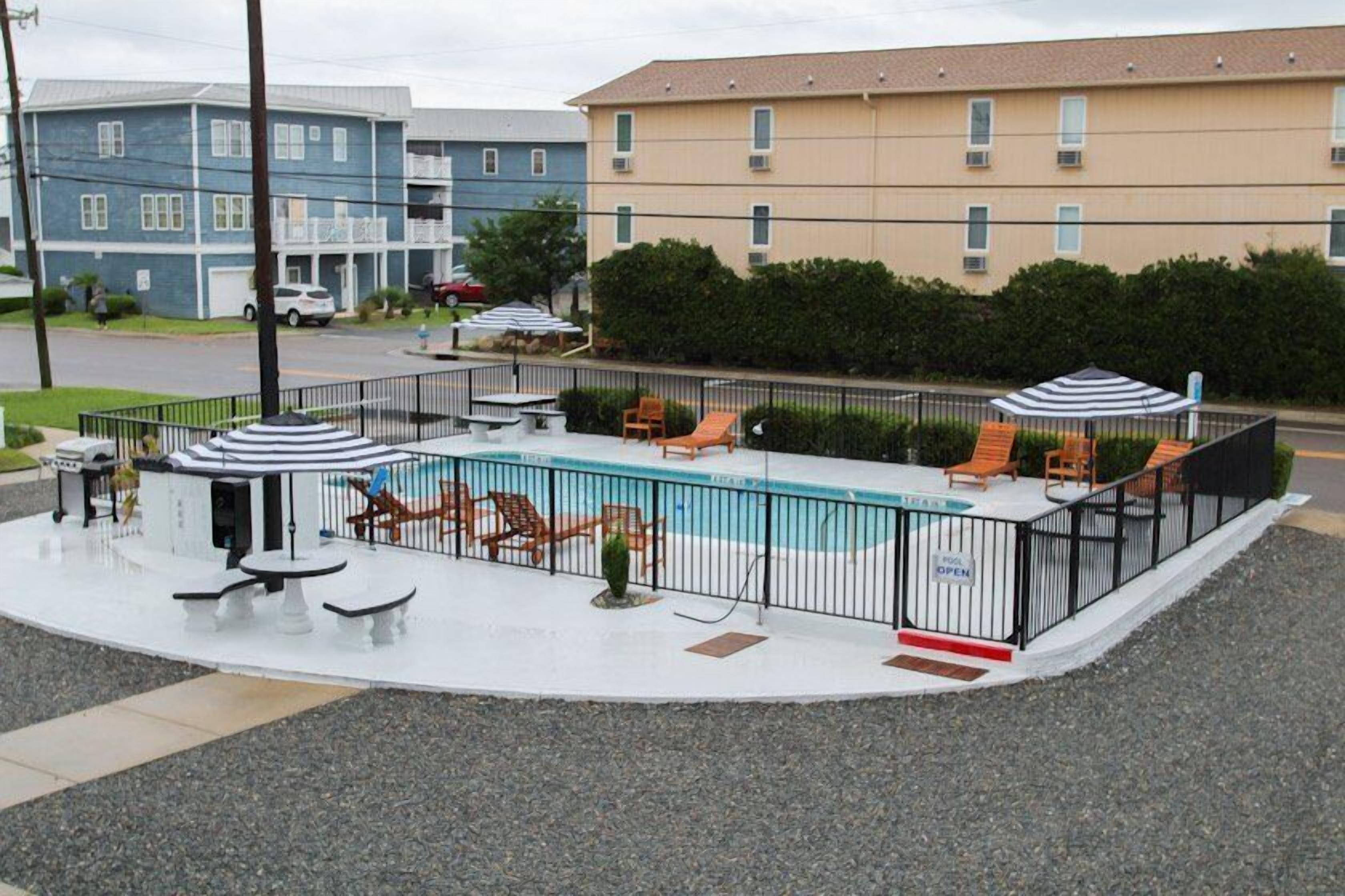 Zona de piscina con sillones de descanso y sombrillas, rodeada por una valla, con edificios al fondo.