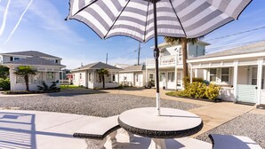 Un patio con una mesa redonda y un paraguas a rayas, rodeado por un barrio residencial con casas y una palmera.