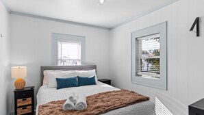 Un dormitorio con cama, dos mesitas de noche, lámpara, ventana con persianas y vistas al exterior.