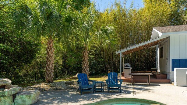 A pool with two blue Adirondack chairs, a small table, and a covered patio area with a white building and blue accents.