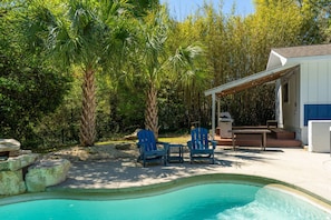 Una piscina con dos sillas Adirondack azules, una mesita y un porche cubierto con un edificio blanco y detalles en azul.