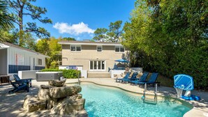 A swimming pool with lounge chairs, a basketball hoop, and a building with a flag.