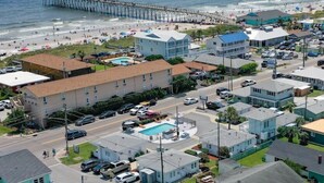 Un pueblo costero con un muelle que se adentra en el océano, una playa con gente y un hotel con piscina y varios pisos.