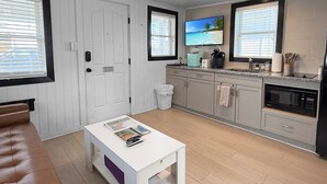 Una cocina moderna con armarios grises, encimera blanca y horno empotrado. Hay una puerta blanca con ventana, un cubo de basura blanco y un sofá de cuero marrón en la sala de estar.