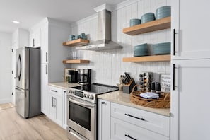A modern kitchen with white cabinetry, stainless steel appliances, and teal dishware on wooden shelves.