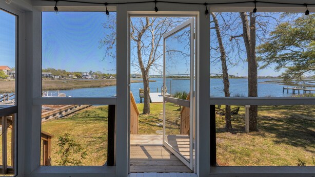 Vista a través de un ventanal grande de una terraza que da a una zona verde junto a un lago, con árboles y un muelle.