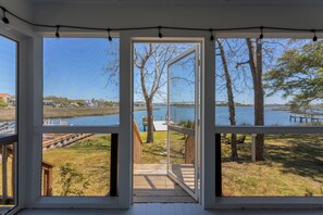 A view through a large window of a deck leading to a grassy area by a lake with trees and a dock.