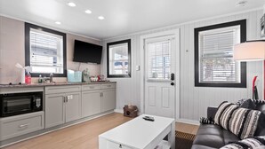 A modern kitchen with white cabinets, a microwave, and a flat-screen TV. There is a white coffee table and a black leather sofa with patterned cushions.