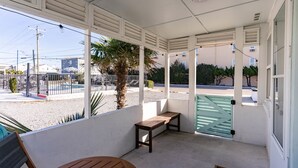 A covered outdoor seating area with a bench and a table, a palm tree, and a view of the street.
