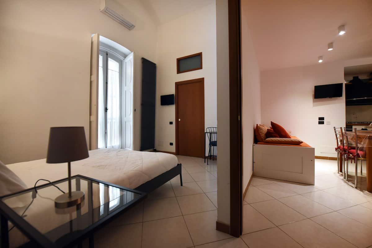 Quarto moderno com mesa de cabeceira de vidro, cama, escrivaninha e um espaço de estar com sofá.