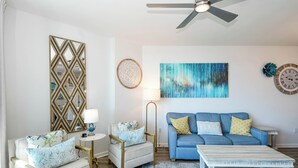 A living room with a blue sofa, a coffee table, a chair, a floor lamp, a ceiling fan, and a large abstract painting.