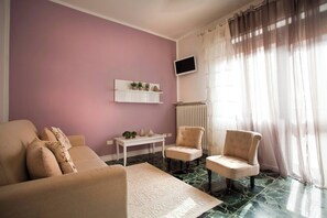 A living room with a sofa, two armchairs, a coffee table, a TV mounted on the wall, and a window with sheer curtains.
