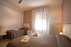 A hotel room with two beds, a bedside table, a chair, a radiator, and a window with sheer curtains.