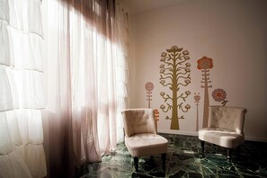 Quarto com duas poltronas, uma janela grande com cortinas leves e um mural de parede com motivos florais e de árvores.