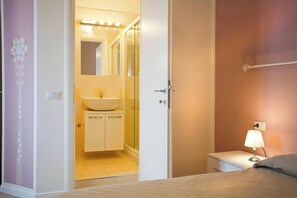 Quarto de hotel com cama, mesa de cabeceira com luminária, banheiro com pia e espelho, e porta que dá para outro cômodo.