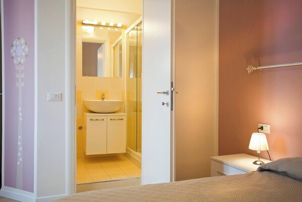 A hotel room with a bed, a bedside table with a lamp, a bathroom with a sink and mirror, and a door leading to another room.